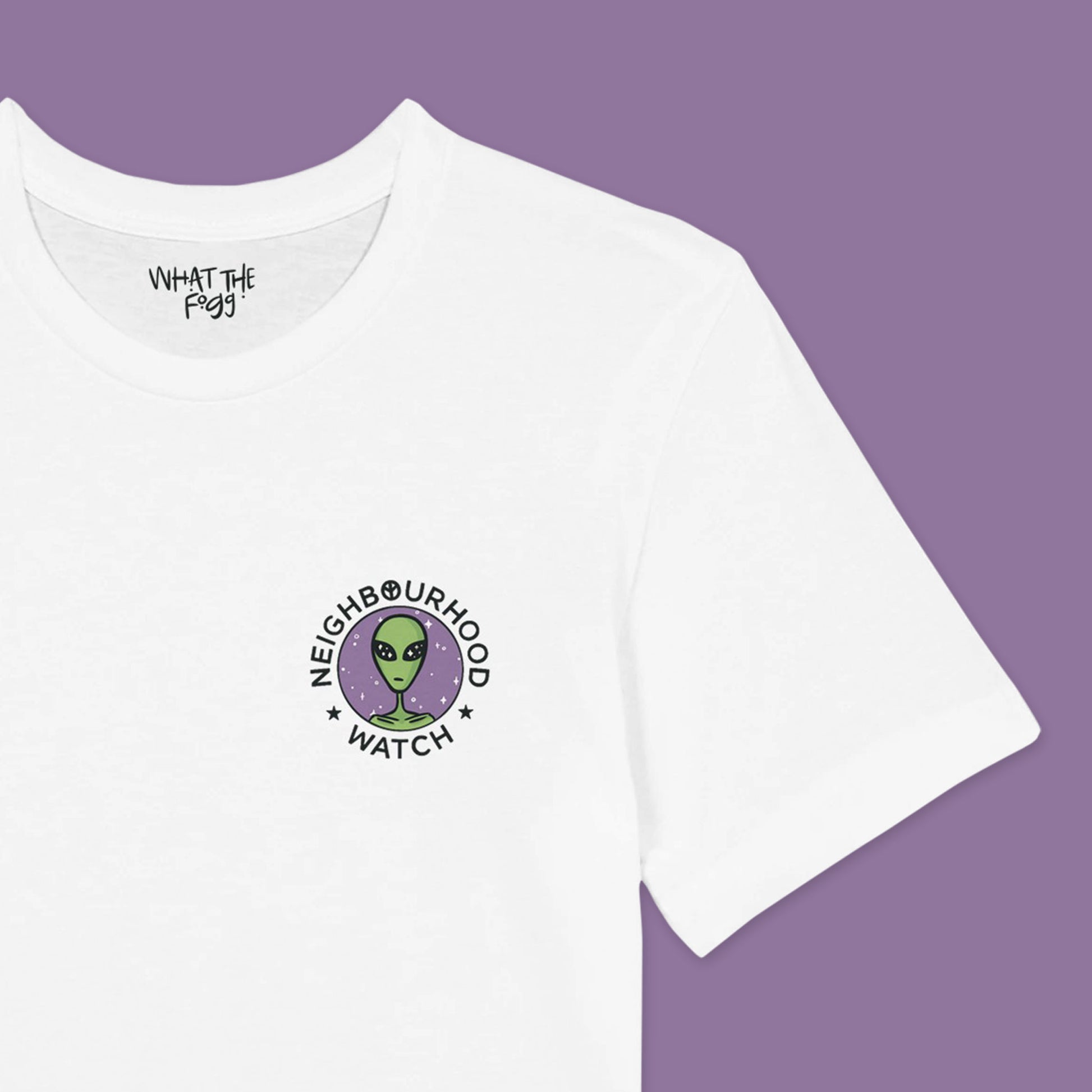 Close up of a White t-shirt featuring a playful twist on the traditional Neighbourhood Watch poster. The front of the shirt displays a circular badge with a design of aliens instead of people. The badge is surrounded by the text 'Neighbourhood Watch' in a bold, eye-catching font.